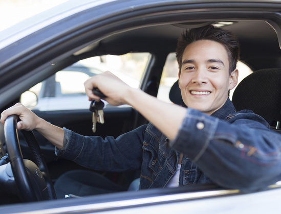 A smiling driver in a denim jacket sitting behind the wheel of a car, holding car keys